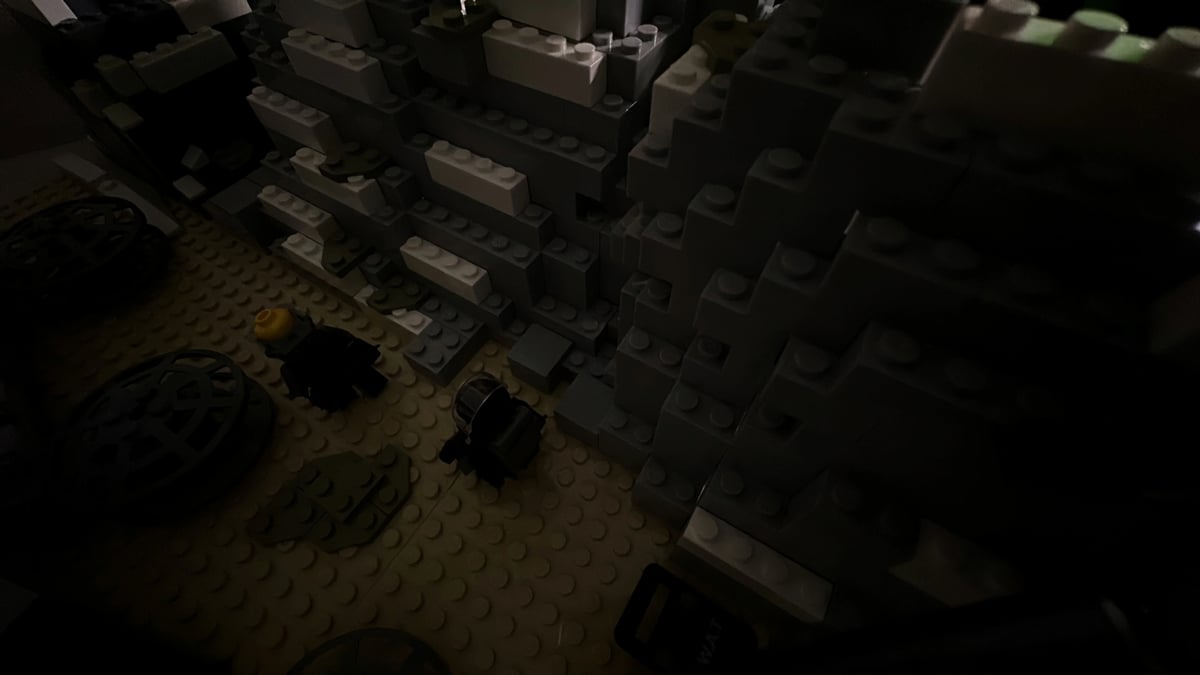 Dim overhead view of a LEGO-like interior room with block walls, floor studs, and small figures and objects on the ground.