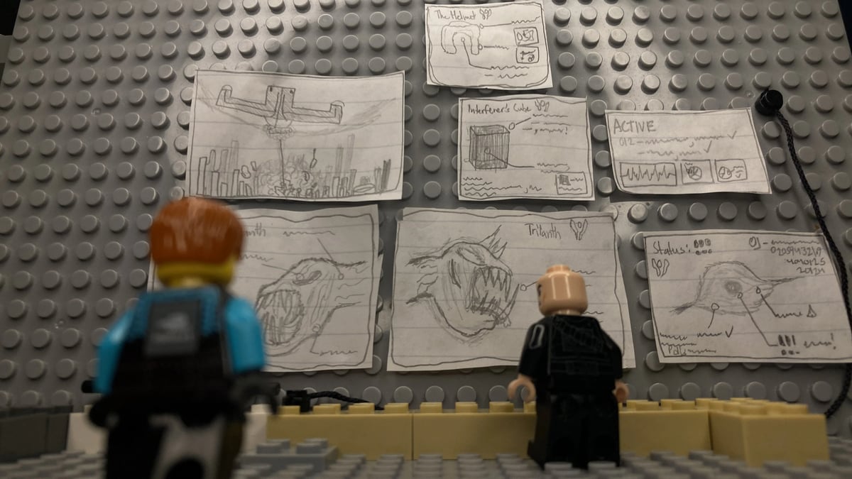 Overhead view of multiple hand-drawn story/creature sketches on paper placed on a gray studded board, with LEGO-like toy figures and blocks at the bottom.