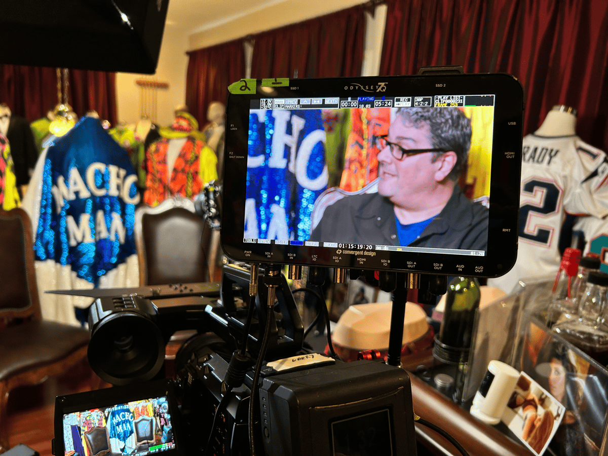 Behind-the-scenes view of a film camera and monitor showing a man being filmed in a room filled with colorful costumes and props
