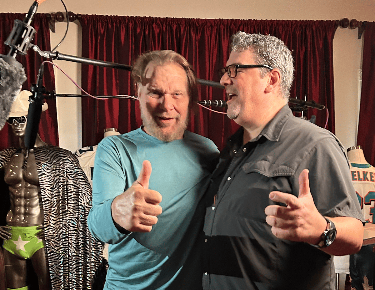 Two men give thumbs-up while speaking beside microphones on a film set with red curtains and theatrical props.