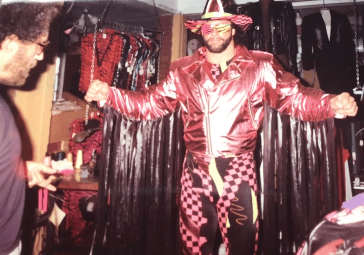Costumed performer in a shiny red jacket, striped hat, and patterned pants poses in a backstage dressing room
