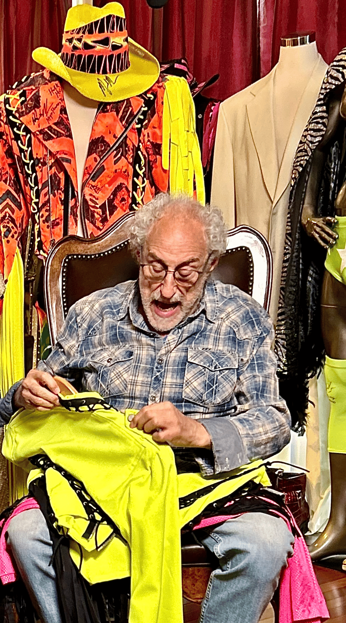 Older man sewing a bright yellow costume in a colorful wardrobe room surrounded by costumes and mannequins
