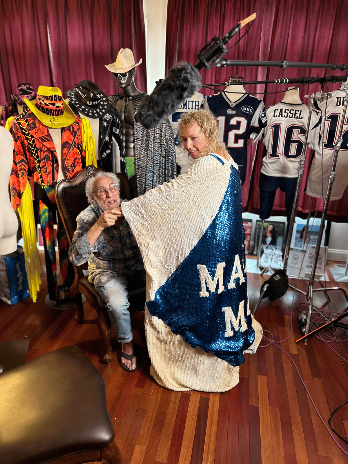 Two people in a costume-filled studio, with colorful garments, mannequins, sports jerseys, and film equipment visible around them.