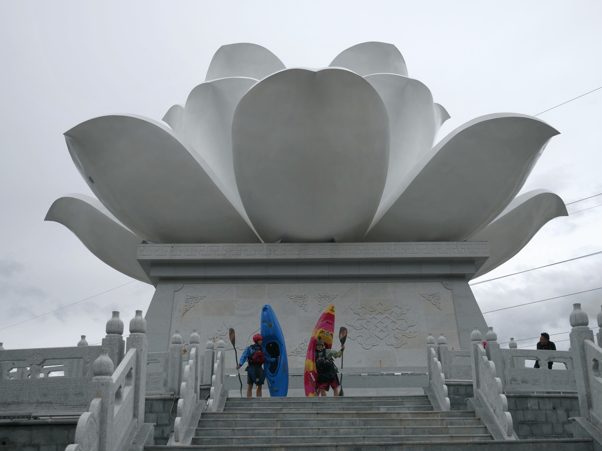 Two colorful figures stand on temple steps beneath a large gray lotus sculpture, with ornate railings and an overcast sky.