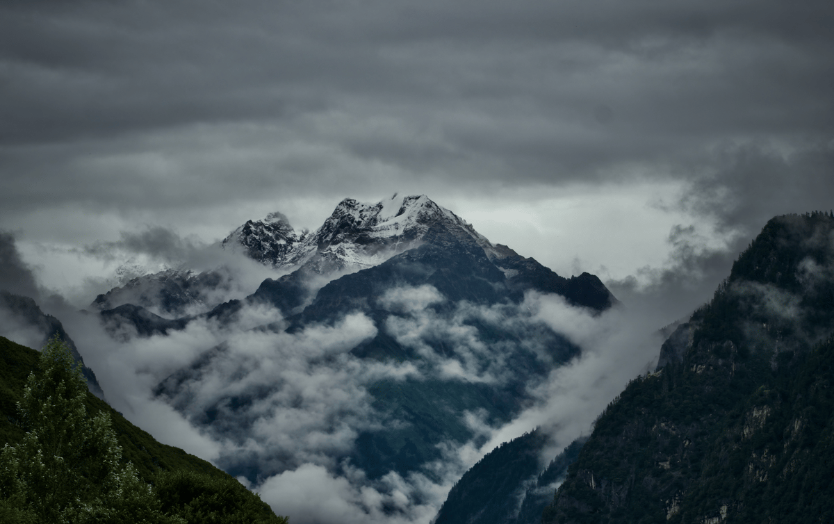 Snow-capped alpine mountains surrounded by swirling clouds and mist beneath a dramatic stormy sky