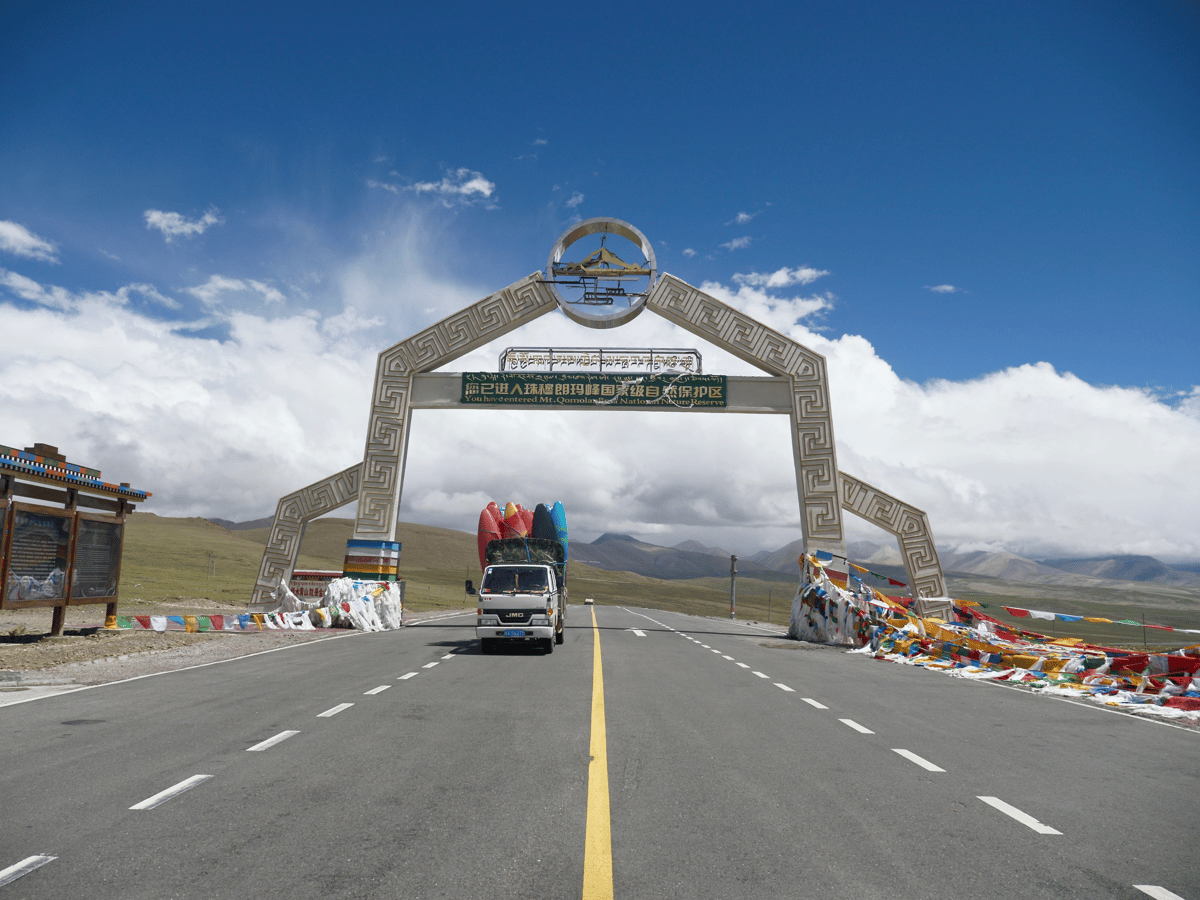 Decorated gateway over a mountain road with a truck approaching, colorful prayer flags, dramatic clouds, and distant rugged peaks.