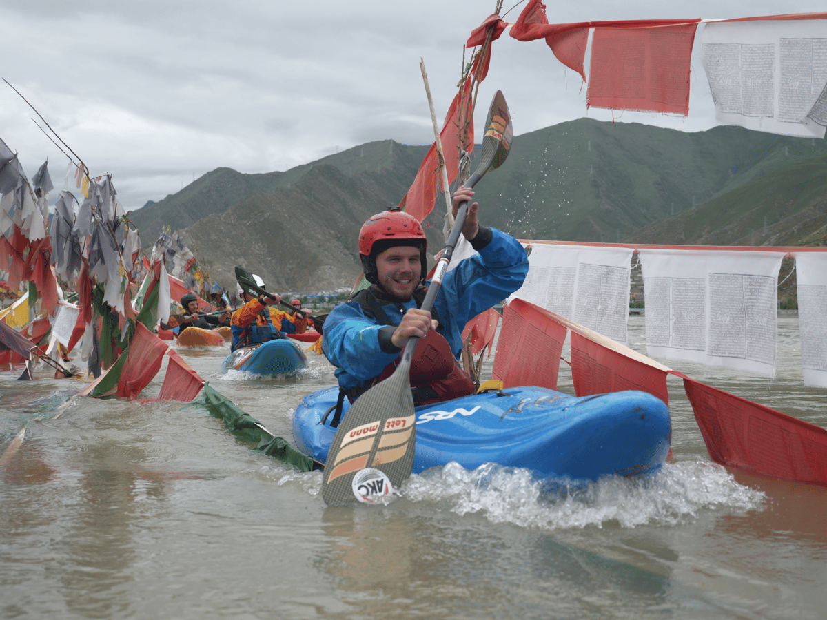 A smiling kayaker in a blue boat paddles through choppy water beneath colorful prayer flags, with green mountains behind him.