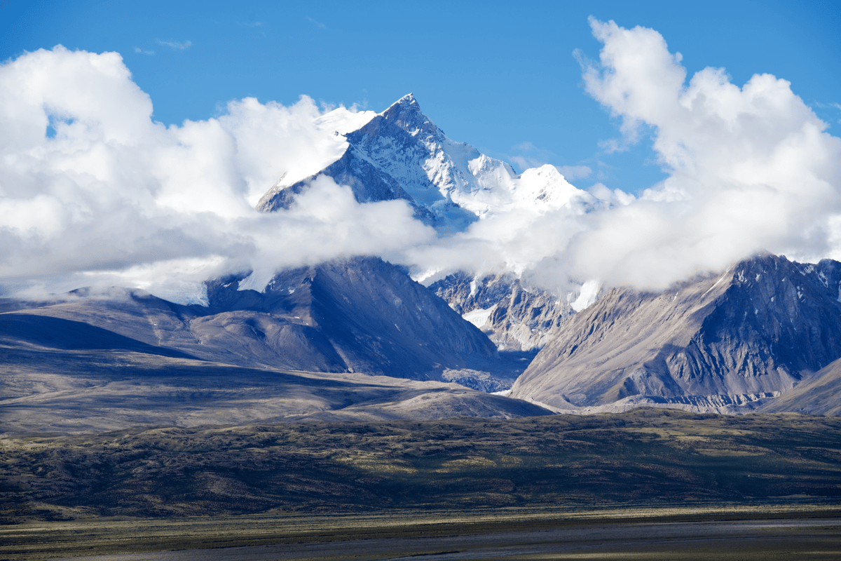 Snow-capped mountain peaks rise above a broad alpine valley, surrounded by dramatic white clouds under a bright blue sky.