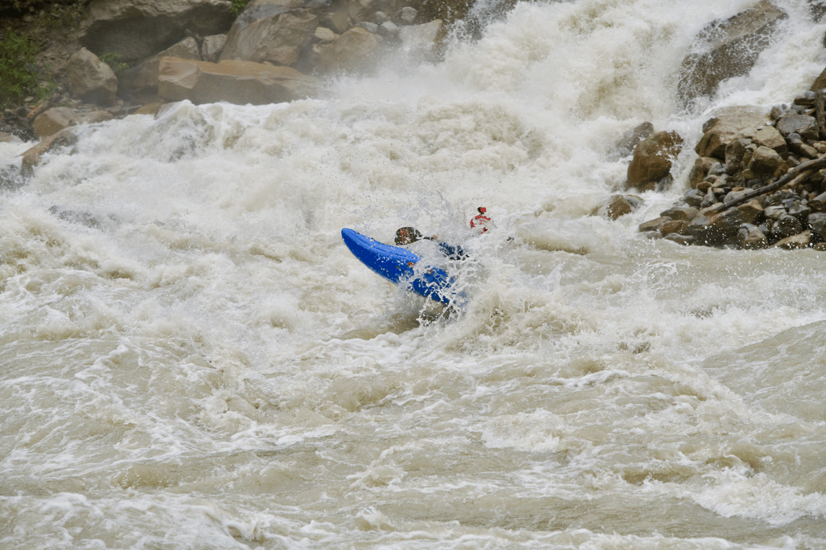 A blue kayak with two visible paddlers moves through powerful whitewater rapids beneath a rocky waterfall.