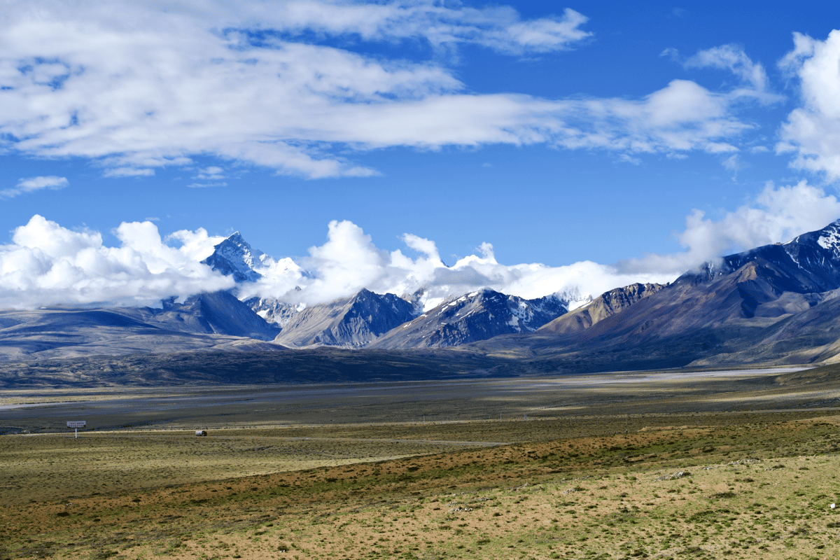 Wide landscape of snow-capped mountains beneath a bright blue sky, overlooking a vast grassy valley.