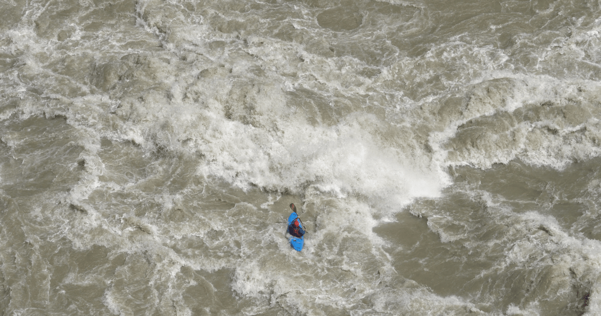 Aerial view of a lone person in blue surrounded by turbulent gray-white water and foamy rapids