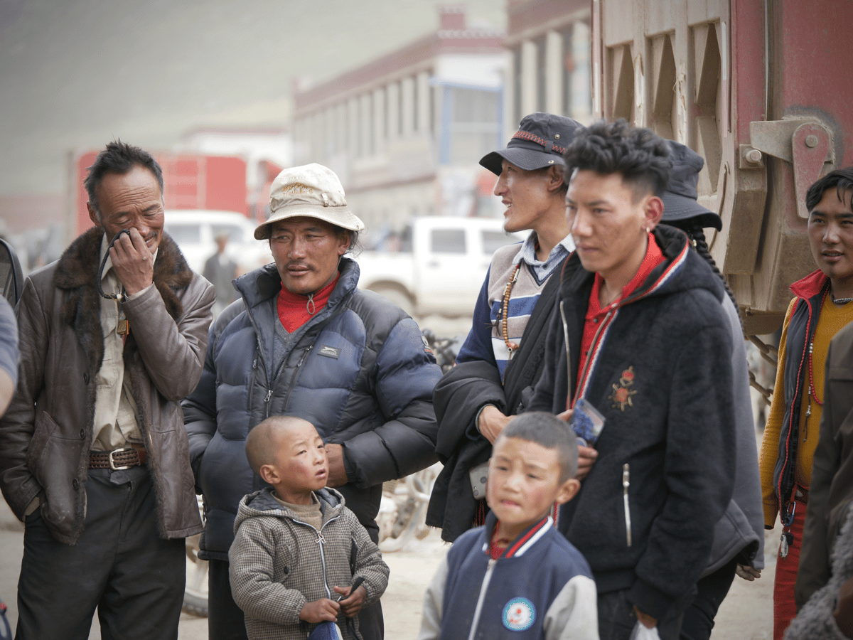 Adults and children gather outdoors on a street, with several people facing the camera in a candid documentary-style scene.