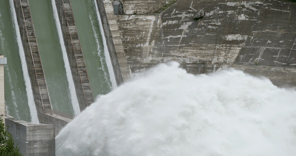 Powerful white water surges from a concrete dam spillway in a wide landscape view.