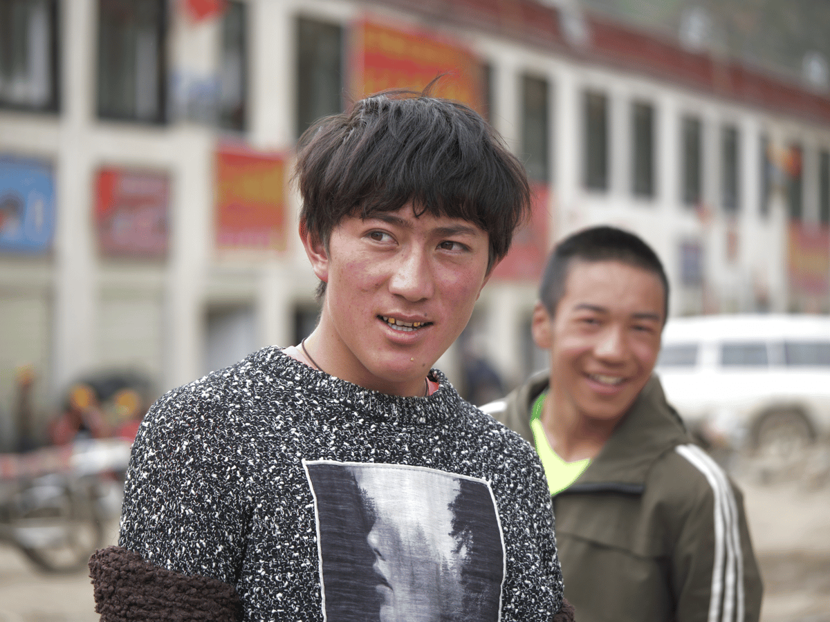Two young men pose outdoors on a street, with one in a speckled sweater in the foreground and the other smiling behind him.