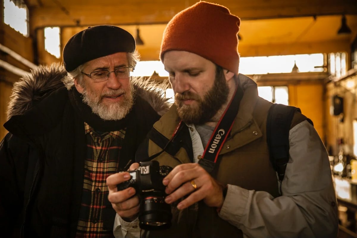 Two bearded men in caps examine a camera together indoors, warm light in the background.