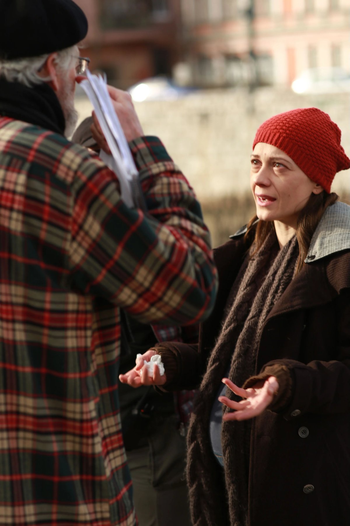 A man in a plaid coat and beanie speaks with a woman wearing a red knit hat and scarf outdoors in winter light.