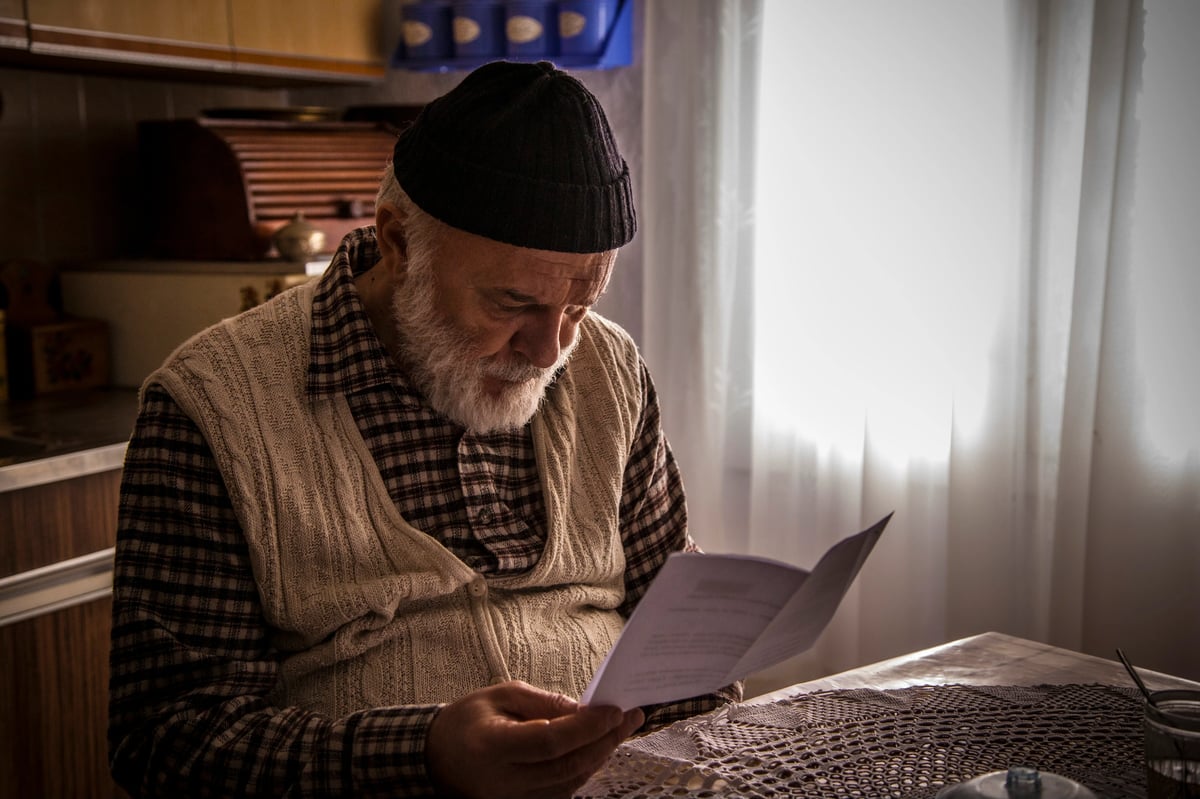An elderly man in a dark knit cap and plaid clothing sits indoors reading from a paper by a bright window.