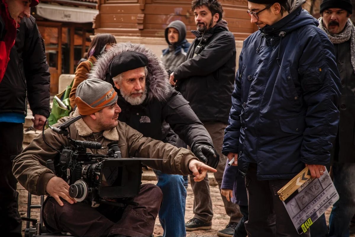 Camera operator on set with winter clothing while crew and an older man stand in the background.