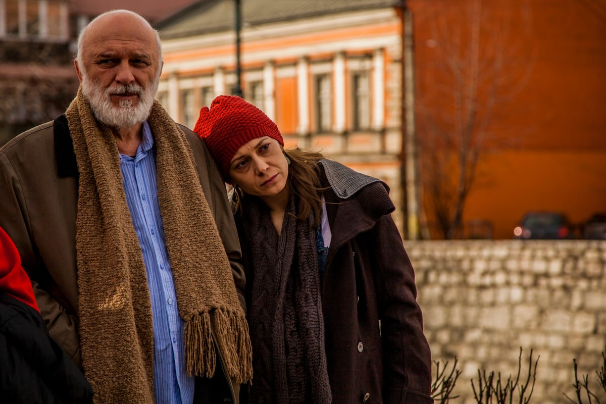 An older man with a gray beard and a woman wearing a red knit hat and winter coats stand outdoors in front of a building.