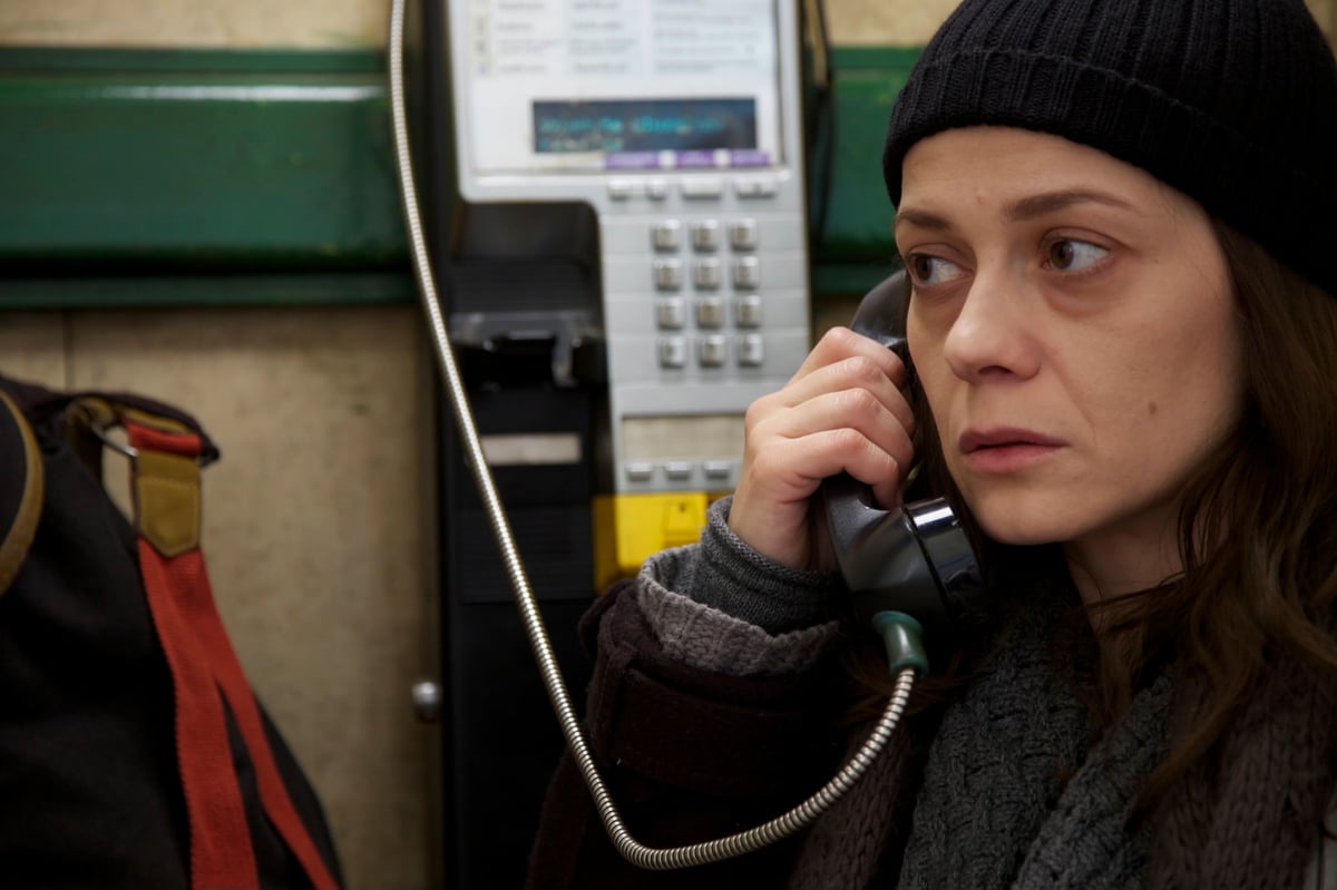 A woman wearing a dark knit hat holds a phone to her ear in a room with blurred computer equipment behind her.
