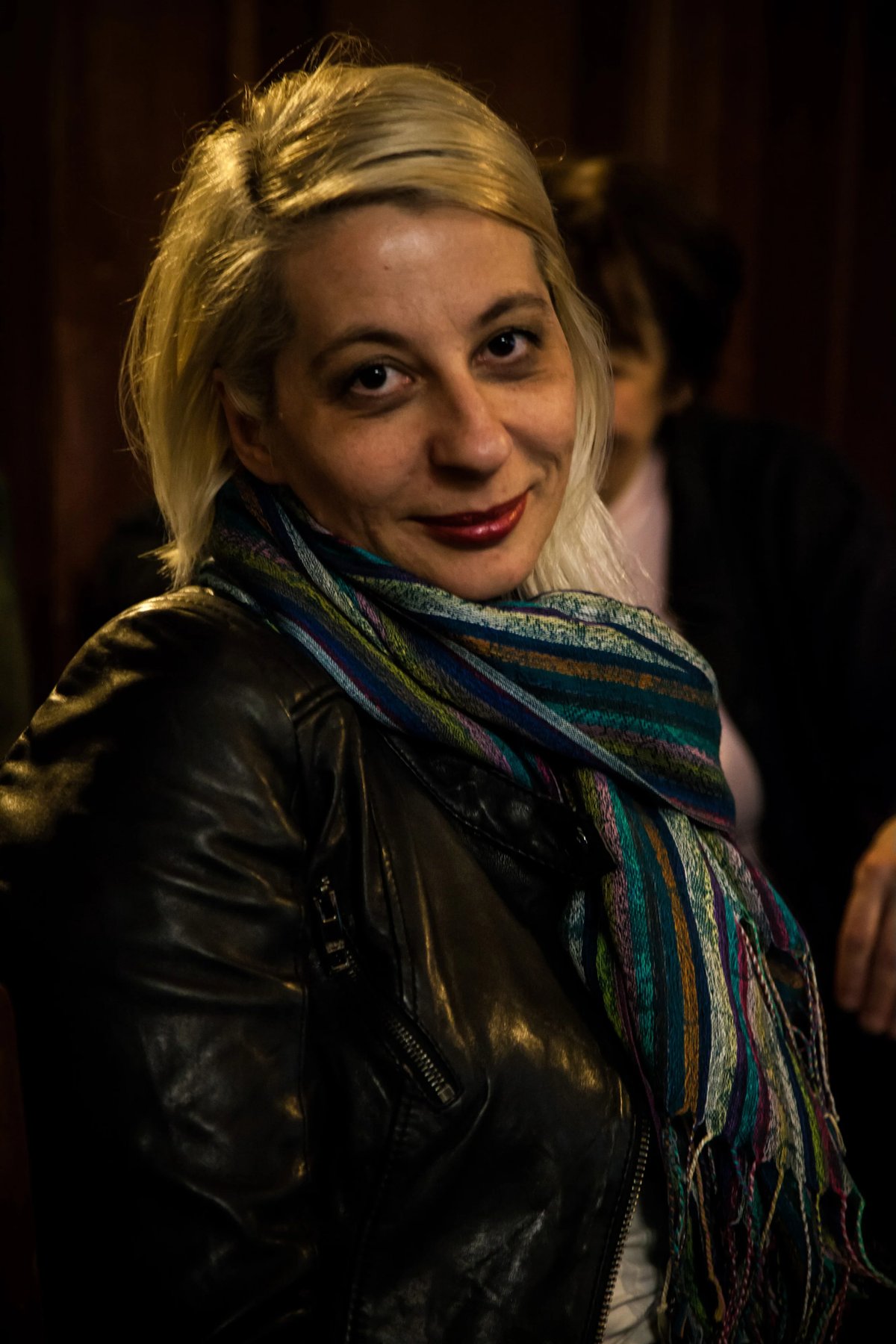 A blonde woman with makeup and a colorful striped scarf smiles in a close-up portrait with a softly blurred background.