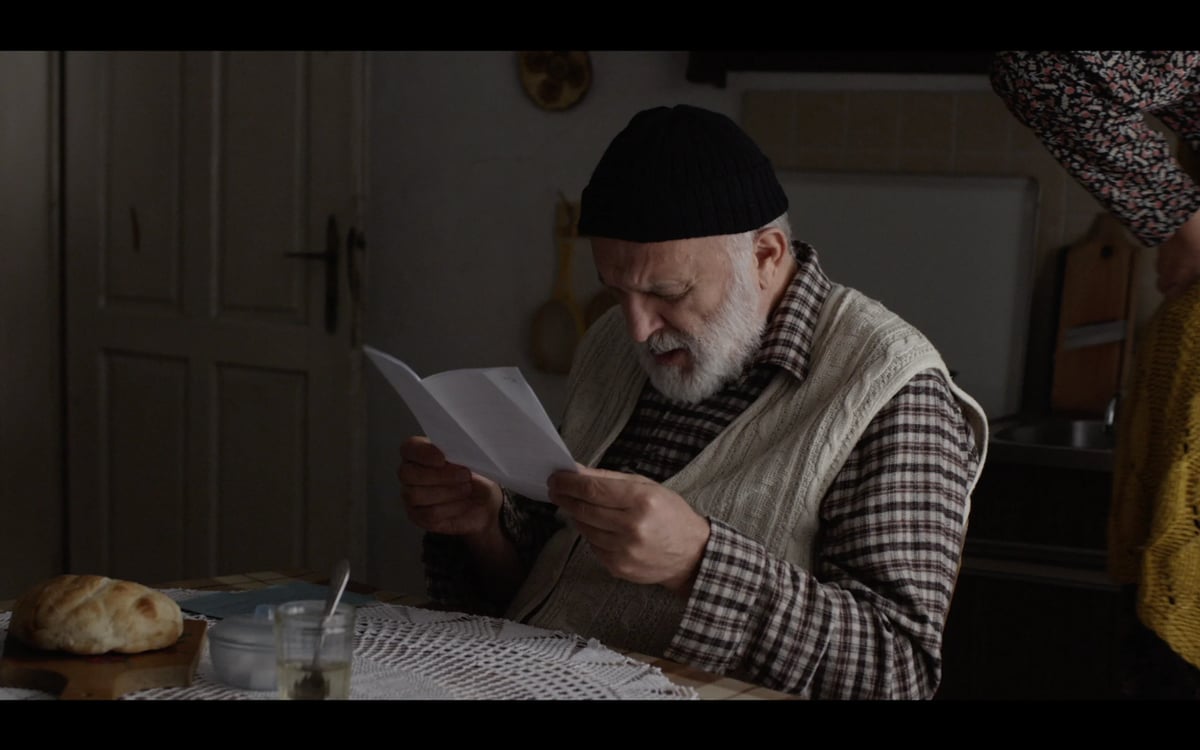 An older man wearing a dark cap reads a document while sitting at a table indoors.