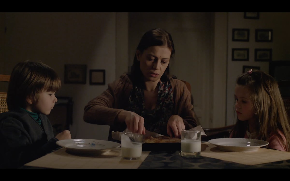 Woman seated at a dining table with two children, looking down at their food in a dim living room setting.
