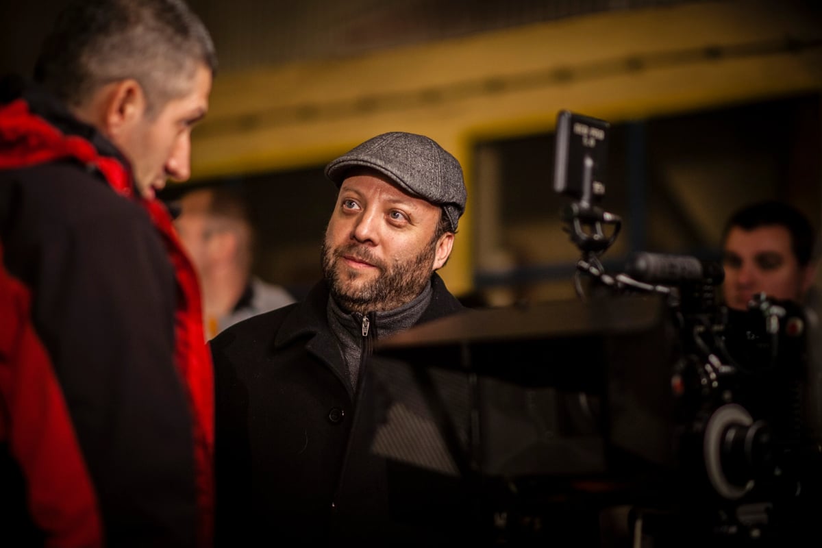 A man wearing a flat cap stands on a film set, looking toward the camera, with another person blurred in the background and a video camera in the foreground.