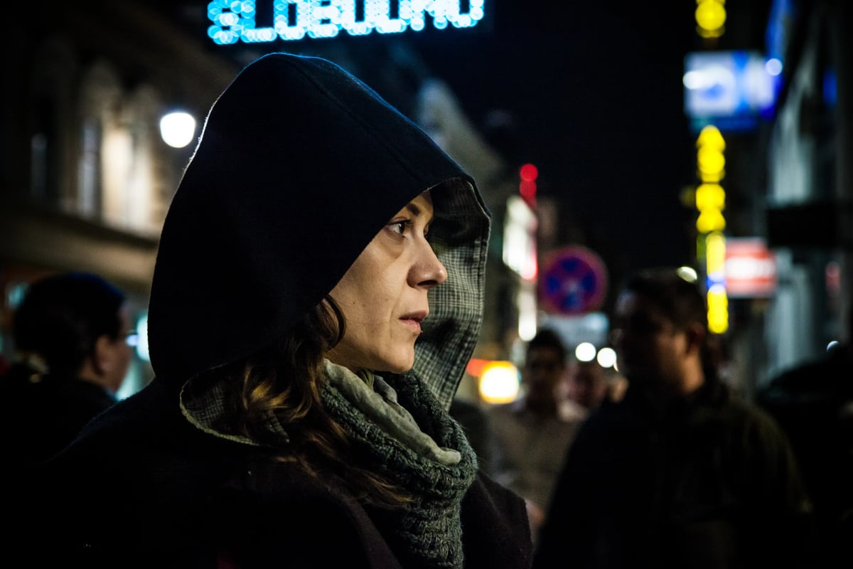 A woman wearing a dark hood and scarf looks to the side in a nighttime street scene with blurred lights and pedestrians in the background.