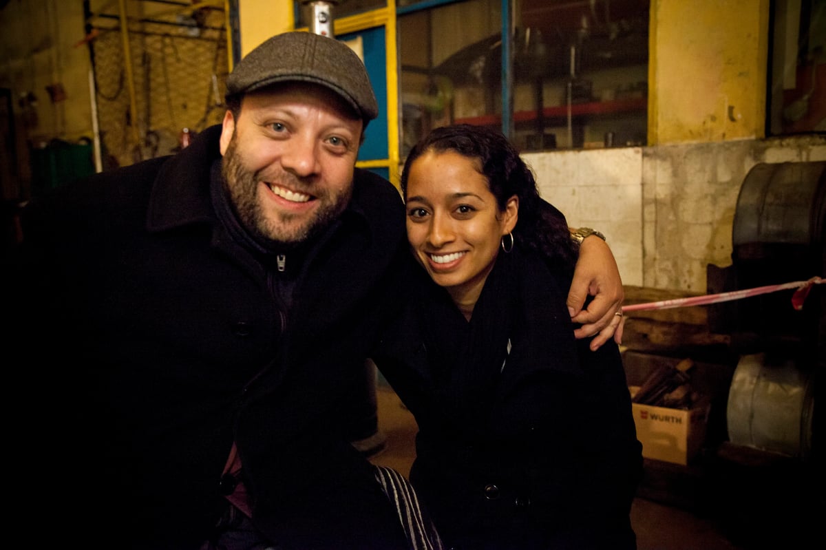A smiling man in a cap and hoodie and a smiling woman in a dark hoodie pose together indoors.