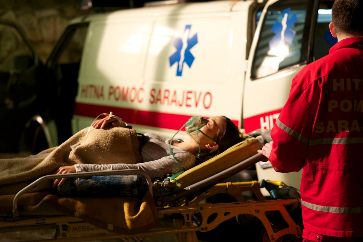 A paramedic in a red jacket tends to a patient on a stretcher near an ambulance while emergency equipment is visible in the scene.