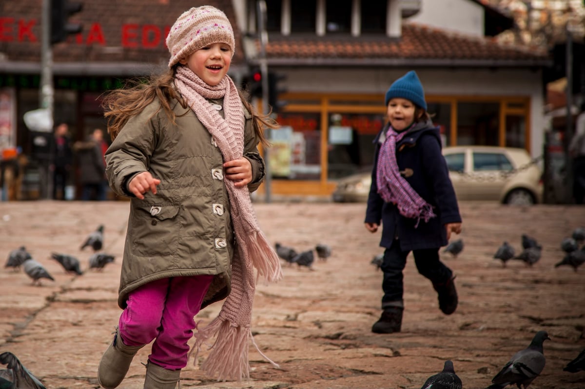 Two children in winter hats and scarves run through a courtyard, with pigeons on the ground and buildings in the background. A second child is visible behind them.