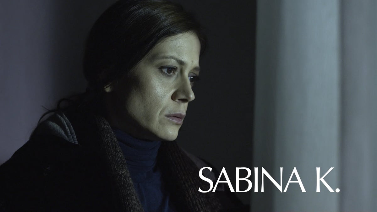 Woman shown in close-up profile under dim lighting, with the text “SABINA K.” displayed at the bottom.