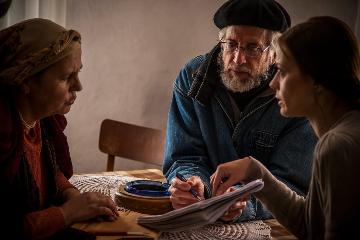 Older man wearing a beret and glasses sits at a table talking with two women, with documents and writing tools in front of them.