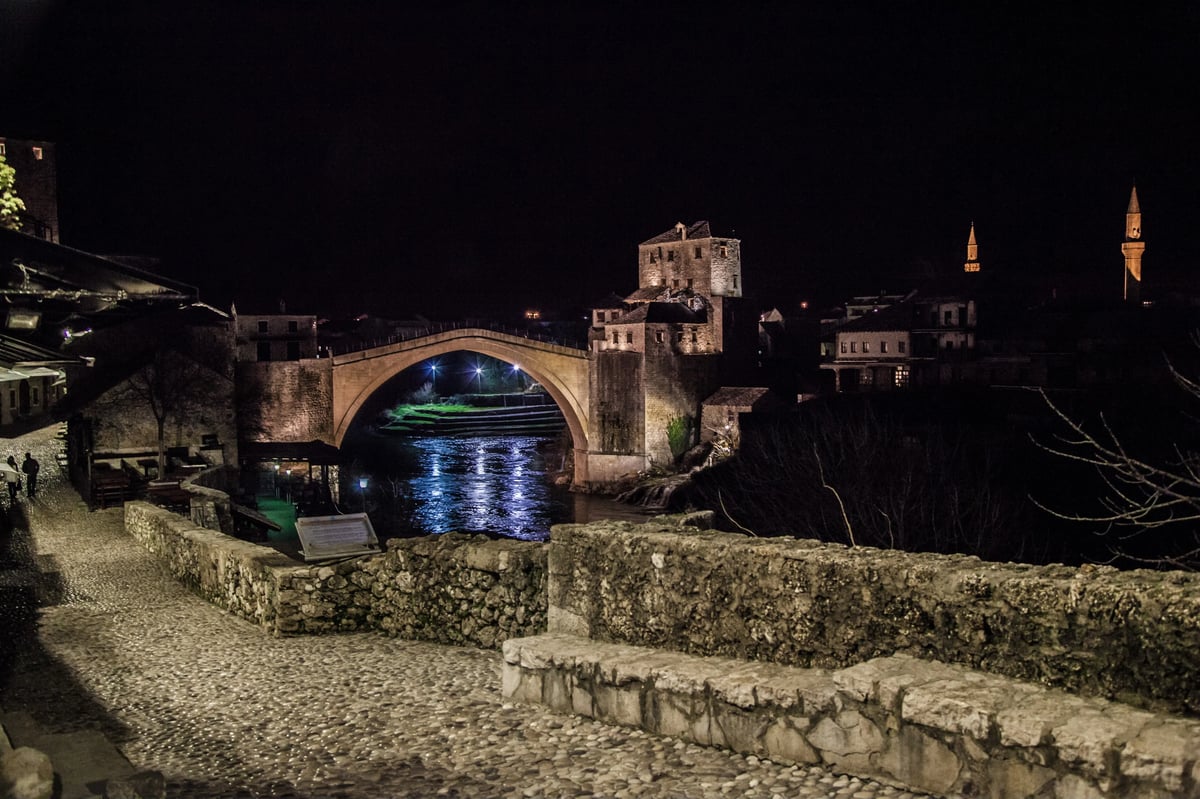 A nighttime view of a lit stone arched bridge over a river in a historic town with buildings on both sides.