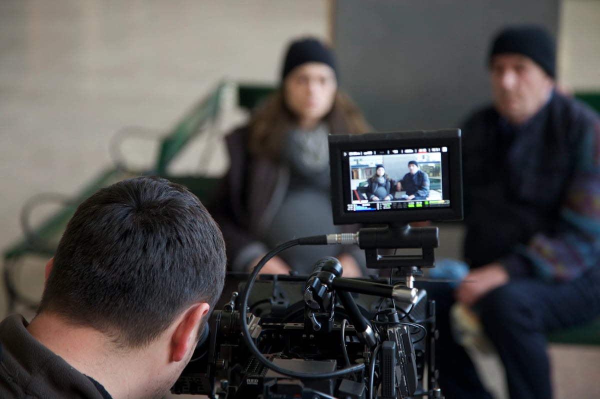 Blurred film crew and actors visible on a camera monitor during a shoot, with a professional cinema camera in the foreground.