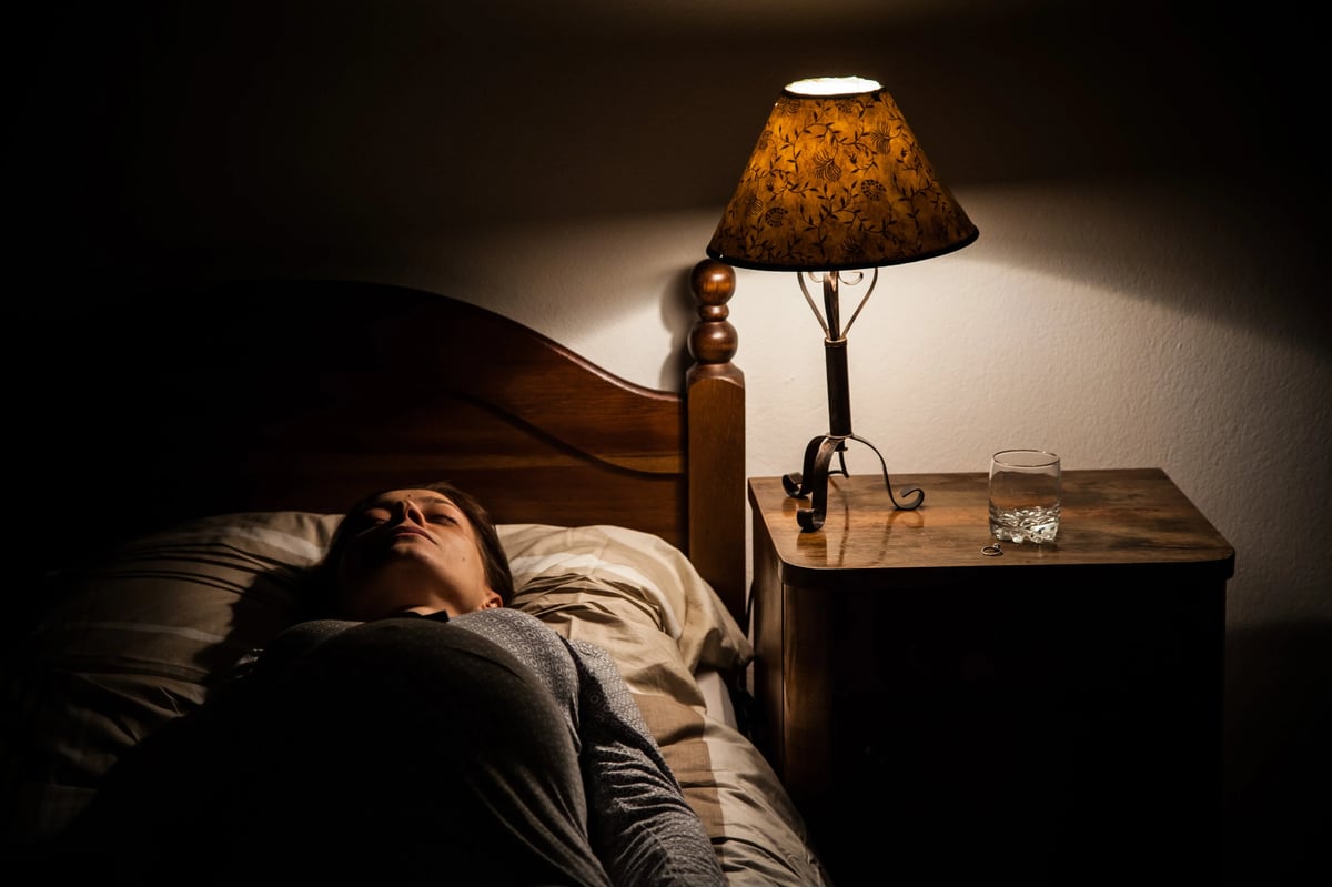 A person lying in bed at night as a bedside lamp casts warm light over a wooden table and a glass of water.
