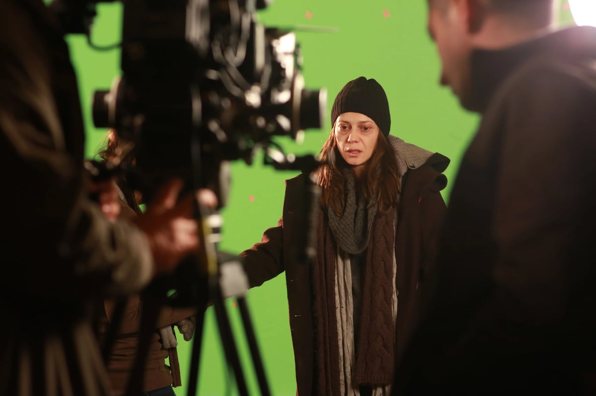 A woman in a beanie and coat stands on a film set in front of a green screen while a camera crew films her.
