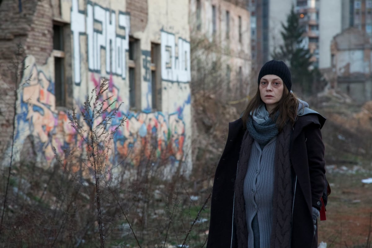 A woman in a dark winter coat and knit beanie stands outdoors beside a colorful graffiti-covered building, looking toward the camera.