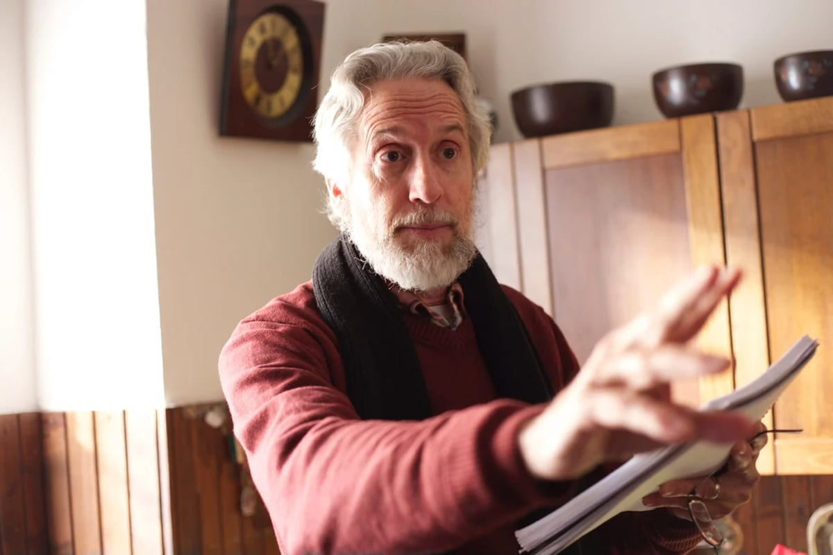 Older man with gray hair and beard reading from papers while gesturing with his hand in a warmly lit room.