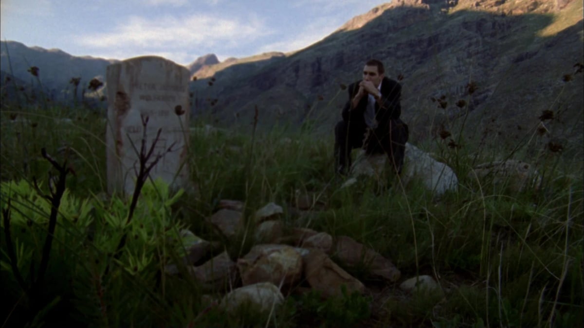 A lone man stands among tall grass in a mountain valley at dusk, with hills and clouds in the distance.