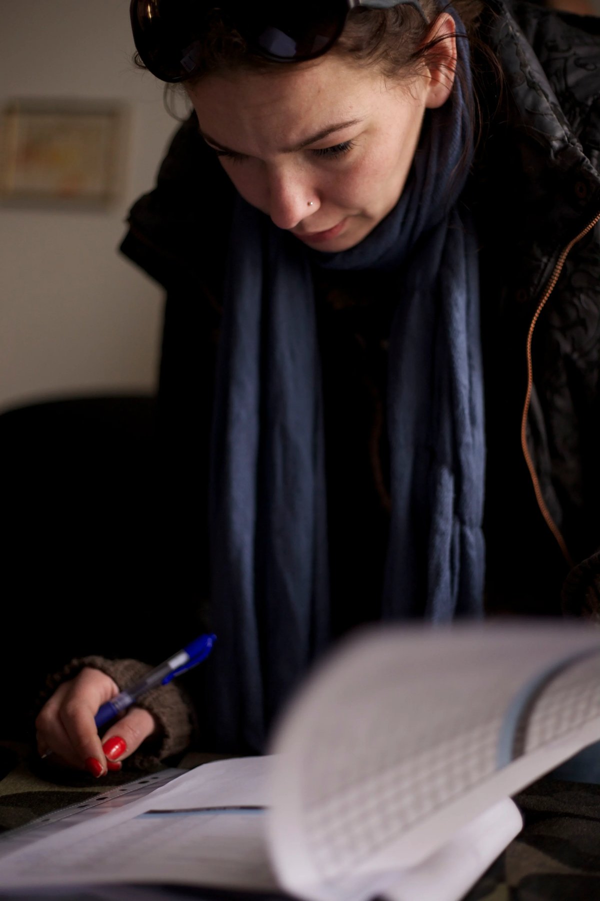 A person in a dark hoodie and sunglasses looks down while writing at a desk with papers.