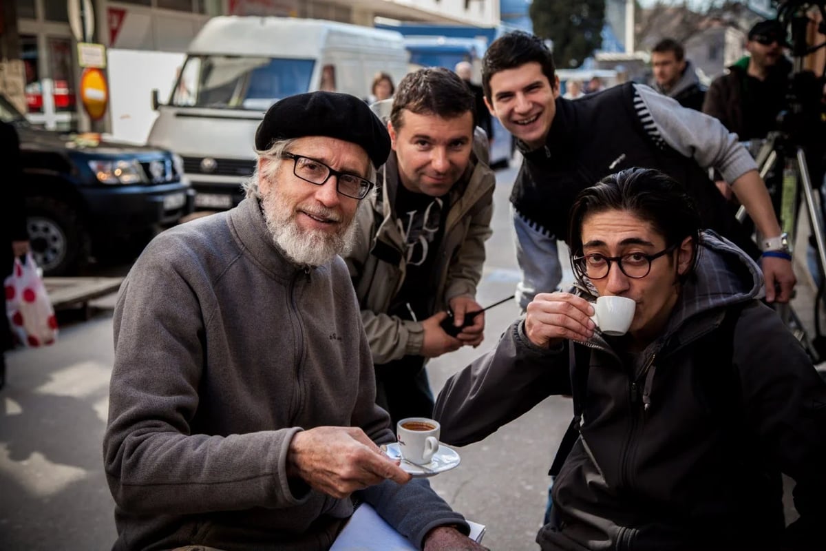 Two people wearing winter coats sit together on a street, one holding a cup and saucer and the other sipping from a white coffee cup, with people and vehicles behind them.