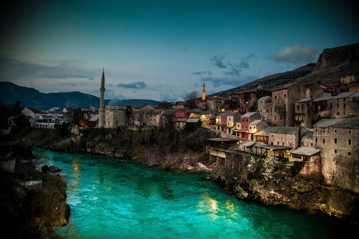 Wide landscape scene of a historic town by a turquoise river at dusk, with mosque minarets, domes, and mountains in the background.