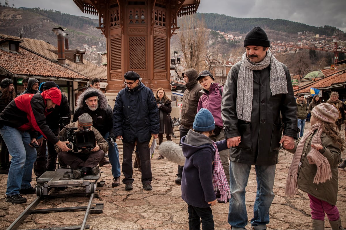 Film crew and cast members in winter coats on a cobblestone street in front of an ornate gate, with a camera rig in the foreground.