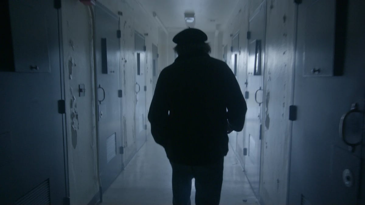 Silhouetted hooded figure in a dark corridor, seen from behind, with bluish lighting and doors on both sides.