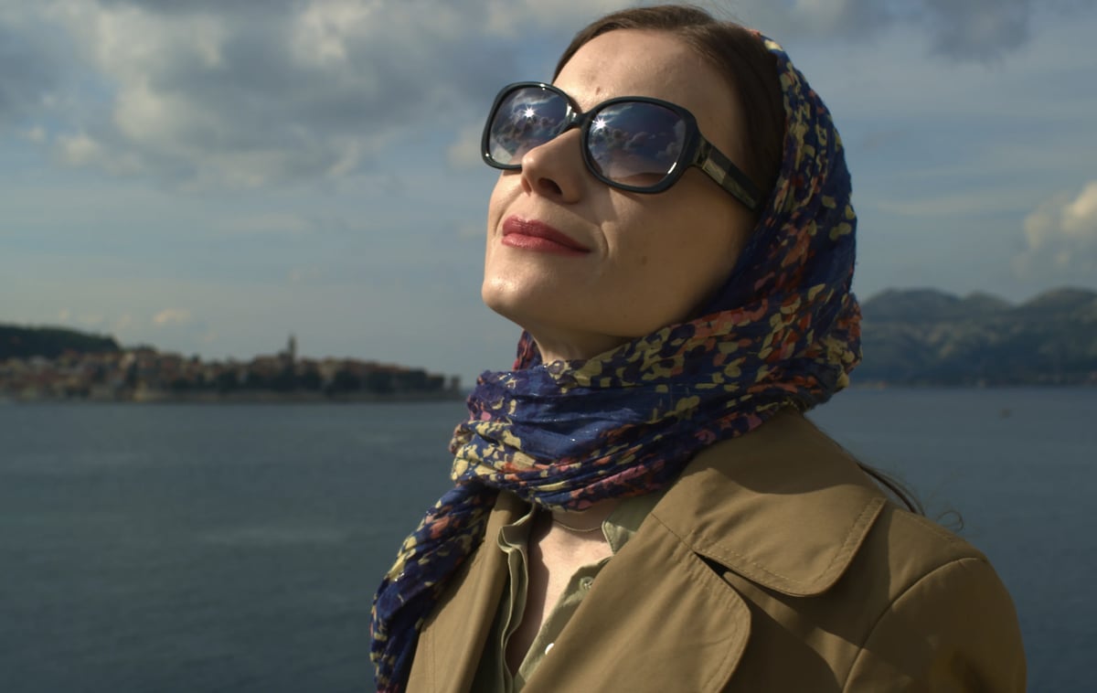 A woman in sunglasses and a patterned headscarf stands outdoors near the water, looking to the side against a blue sky.