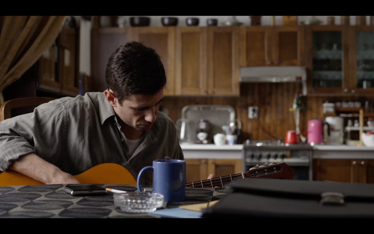 A man in a kitchen looks down while playing an acoustic guitar at a table.