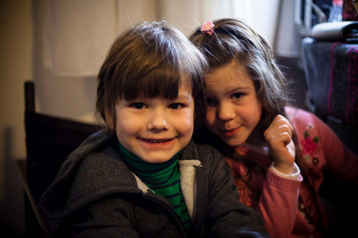 Two children smiling while sitting close together indoors, one in the foreground and one slightly behind.