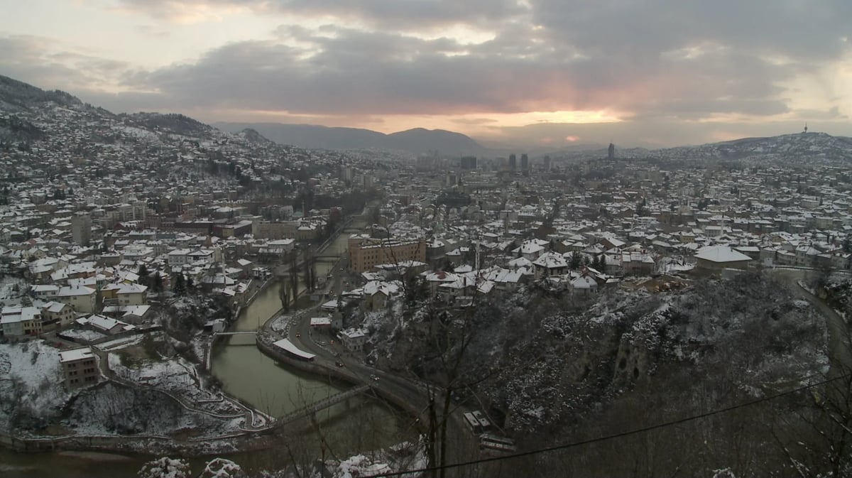 Aerial view of a snowy city beside a winding river under a cloudy dusk sky.